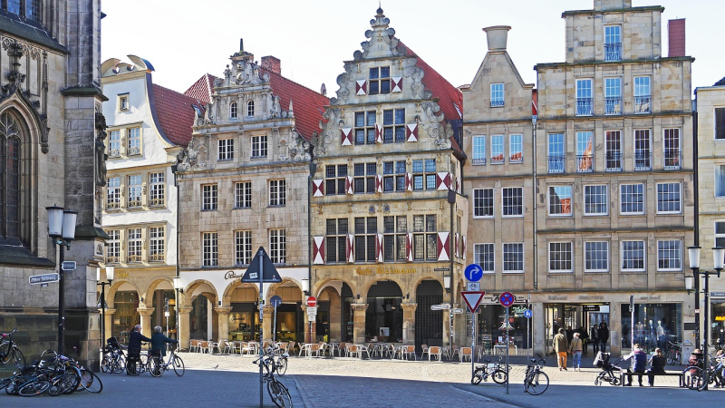 Historische Giebelhäuser am Prinzipalmarkt in Münster, mit roten Dächern und weißen Fensterläden, Fahrräder und Menschen.